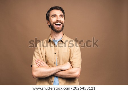 Happy young bearded man. Portrait of handsome young man in casual keeping arms crossed and smiling while standing against beige background.
