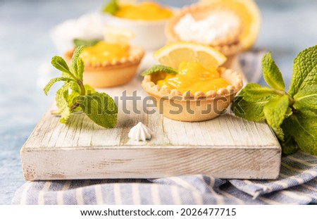 Shortbread tartlet filled with lemon curd, mint and lemon slices and mini meringue on wooden board, blue concrete background. Selective focus.