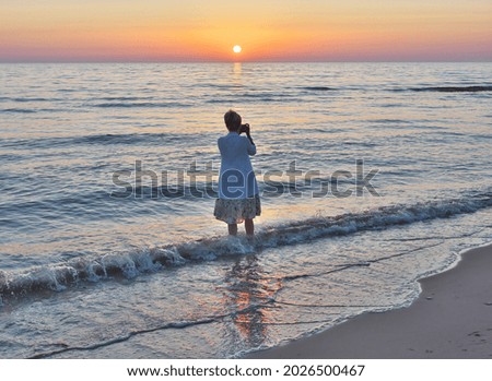 A woman stands in the water and photographs the sunset.