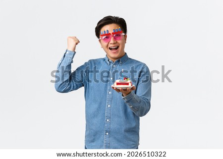 Celebration, holidays and lifestyle concept. Upbeat positive asian guy in funny party sunglasses holding birthday cake and fist pump in hooray gesture, determined bday wish come true