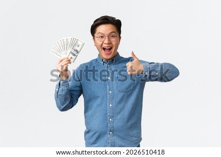 Excited smiling asian man with braces and glasses, showing thumbs-up and waving money, receive paycheck, freelancer got job with good payment, holding cash and looking satisfied