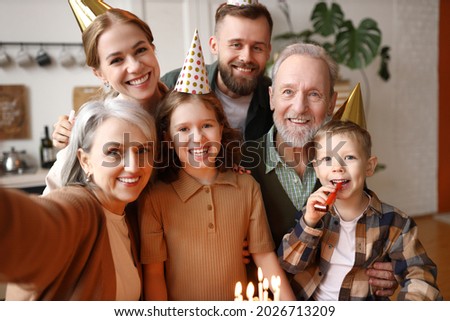 Happy positive big caucasian family grandparents, parents and two kids making selfie during birthday celebration at home, wearing party hats and smiling cheerfully at camera while standing indoors