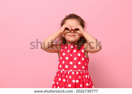 Smiling preschool girl in a pink polka dot dress, imitating a binocular with her hands and looking through it. Adorable child expressing positive emotions posing on pink background with copy space