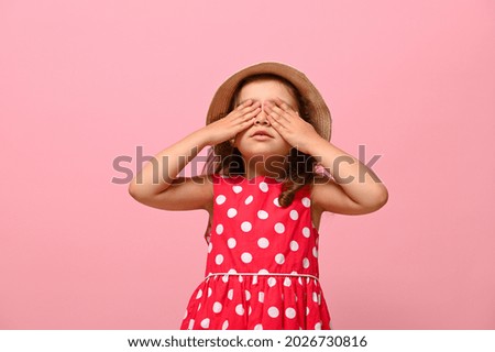 Confident portrait of gorgeous baby girl dressed in summer polka dots dress and straw hat , covering her eyes with her hands, posing over pink background with space for text for advertising