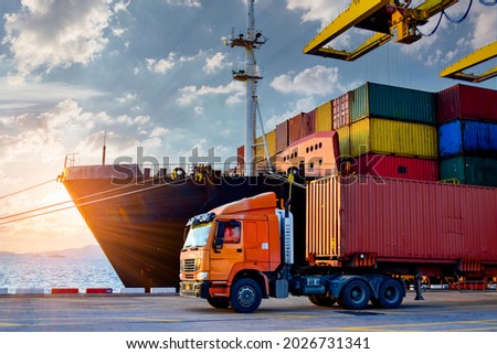 Truck carrying forty-foot container leaving port terminal with ship and quay crane on the background. Seaport operation activities, container shipping, and logistics. Royalty-Free Stock Photo #2026731341