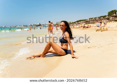 A young tanned girl in a blue swimsuit is laughing cheerfully, sitting on the seashore, blurred background