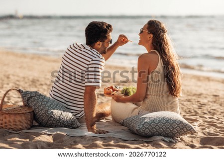 leisure, relationships and people concept - happy couple with food eating and having picnic on beach Royalty-Free Stock Photo #2026770122