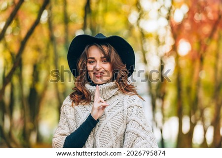 Beautiful woman in a knitted cardigan and hat touches collar by her hands in the park on the background of the autumn landscape