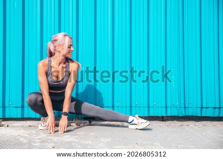 Fitness smiling woman in grey sports clothing with pink hair. Young beautiful model with perfect body.Female posing in the street near blue wall.Cheerful and happy. Stretching out before training