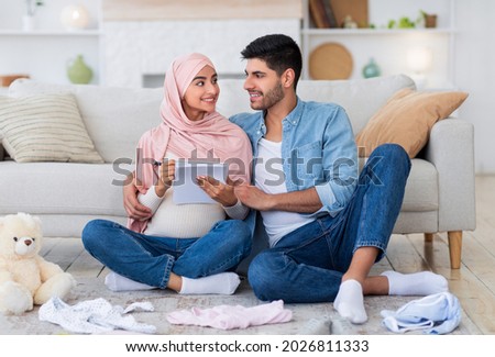 Young pregnant islamic couple getting ready for childbirth, making checklist of necessities while sitting on floor in living room with baby clothes around