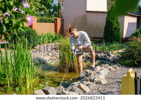 Young smiling caucasian man cleans artificial garden pond bottom with high-pressure washer nozzle from mud and sludge. Spring and summer pond care work