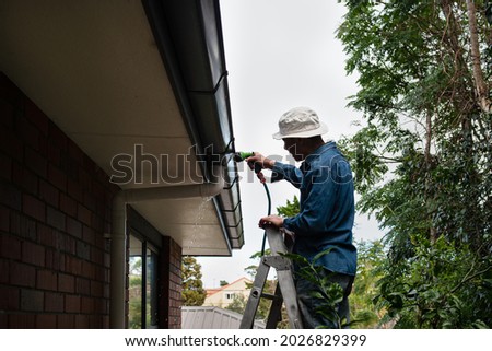 Man standing on the ladder and washing the gutter using a garden hose. Home maintenance work.  Royalty-Free Stock Photo #2026829399