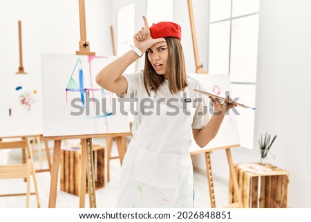 Young artist woman painting on a canvas at art studio making fun of people with fingers on forehead doing loser gesture mocking and insulting. 