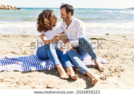 Middle age hispanic couple make selfie by the smartphone sitting on the towel at the beach.
