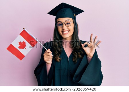 Young hispanic woman wearing graduation uniform holding canada flag doing ok sign with fingers, smiling friendly gesturing excellent symbol  Royalty-Free Stock Photo #2026859420