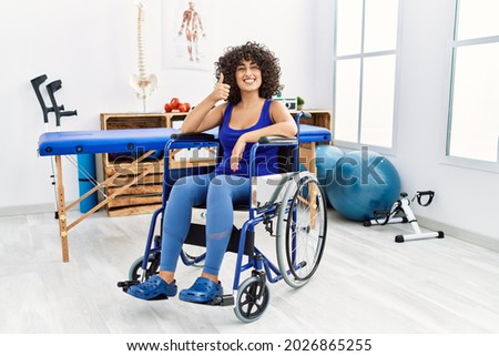 Young middle eastern woman sitting on wheelchair at physiotherapy clinic doing happy thumbs up gesture with hand. approving expression looking at the camera showing success. 