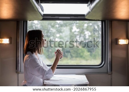 A young beautiful woman in a white shirt is sitting in a train compartment with a Cup of tea and looking out the window. Travel by train.	
 Royalty-Free Stock Photo #2026867955