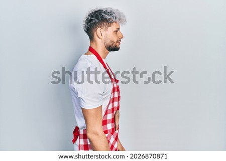Young hispanic man with modern dyed hair wearing apron looking to side, relax profile pose with natural face with confident smile. 