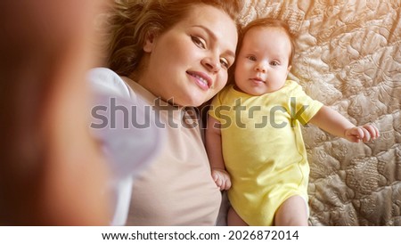 Jolly young mother with plump lips takes picture of smiling baby daughter in yellow with smartphone on bed in apartment room closeup