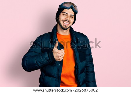 Young hispanic man with beard wearing snow wear and sky glasses doing happy thumbs up gesture with hand. approving expression looking at the camera showing success. 