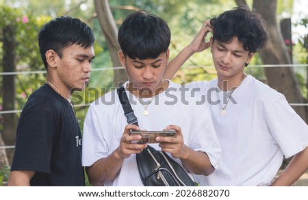 A young man plays online games on his cellphone while two of his friends watch on. One of them is unimpressed at his skill.