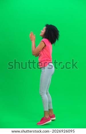 Portrait of young female African American keeping palm together and asking for something. Black woman with curly hair in pink tshirt poses on green screen in the studio. Side view. Full length.