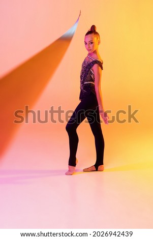 Flying colored ribbon. Portrait of little girl, rhythmic gymnastics artist isolated on pink yellow studio background in neon. Concept of sport, action, aspiration, education, active lifestyle.