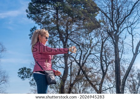 Full side view of an adult woman with sunglasses taking a picture with one hand in a park with blue sky and trees behind.