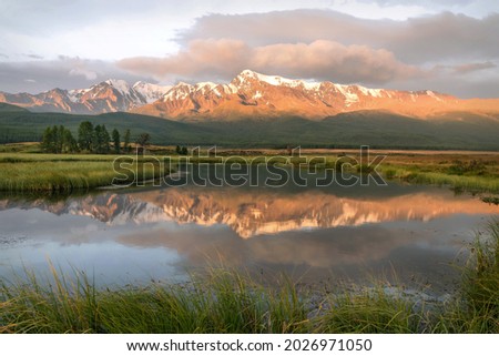 Amazing bright sunrise with mountains covered with snow and forest, green trees, grass, clouds and beautiful mirror reflections in the water. Altai, Russia