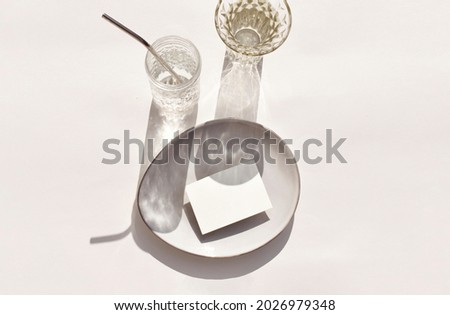 Luxury mockup still life table setting scene. Empty paper blank on stoneware plate, cocktail glass on sunlit background. Mock invitation card for wedding, business or menu. Eco minimalist flat lay Royalty-Free Stock Photo #2026979348