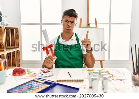 Young hispanic man at art studio holding degree pointing up looking sad and upset, indicating direction with fingers, unhappy and depressed. 