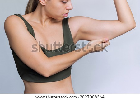 A young caucasian blonde woman  grabbing skin on her upper arm with excess fat isolated on a white background. Pinching the loose and saggy muscles. Overweight concept Royalty-Free Stock Photo #2027008364