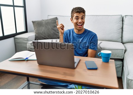 Young handsome hispanic man using laptop sitting on the floor with a big smile on face, pointing with hand and finger to the side looking at the camera. 