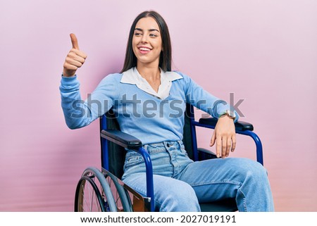 Beautiful woman with blue eyes sitting on wheelchair looking proud, smiling doing thumbs up gesture to the side 