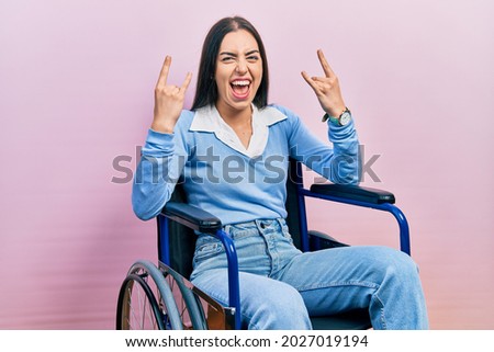 Beautiful woman with blue eyes sitting on wheelchair shouting with crazy expression doing rock symbol with hands up. music star. heavy music concept. 