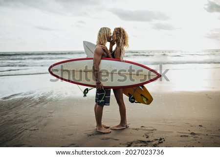 Handsome man and his girlfriend are kissing against background of sea and holding surfboards