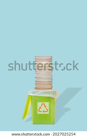Cover page with green garbage trash bin with bag and a pile of paperback books with its shadow and recycling symbol at blue solid background with copy space. Concept of paper recycling