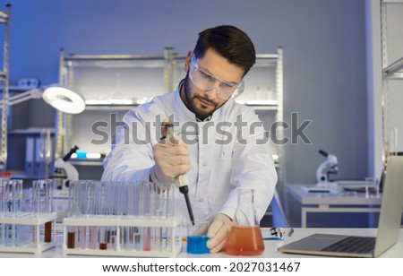 Serious concentrated young male medical scientist, pharma chemist, biotech company employee, lab assistant or student in coat and goggles working alone in science laboratory late in the evening