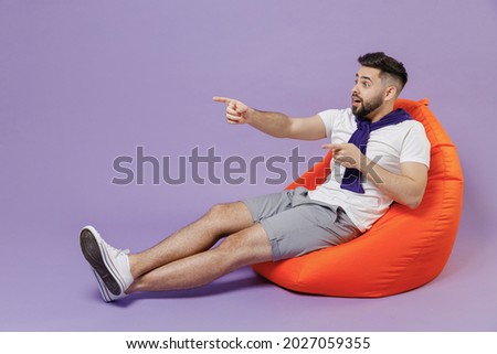 Full size body length young brunet man 20s wear white t-shirt purple shirt sit in bag chair point look aside on workspace area copy space mock up isolated on pastel violet background studio portrait