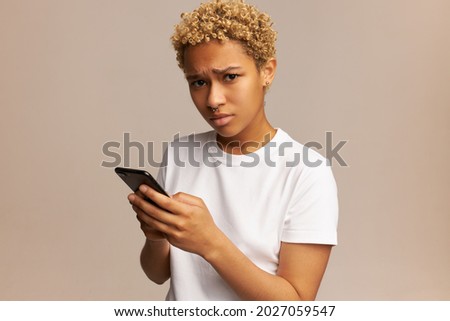 Picture of dark-skinned female student having gloomy, sad face after receiving home task for tomorrow on her smartphone, isolated on pale pink background. People, emotions and feelings concept