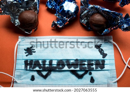 Halloween word written on a medical mask  and some chocolates in their wrappers on a orange surface