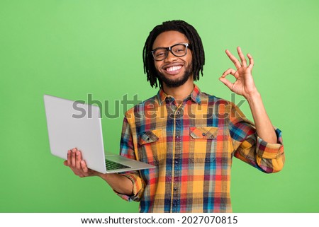 Photo of excited sweet dark skin man wear plaid shirt spectacles showing okey holding modern device isolated green color background