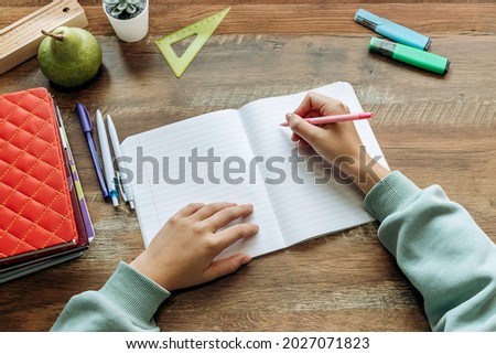 Schoolchild  writing in a notebook at home at his desk.Completing homework.Stationery, textbooks and notebooks are on the table.Back to school concept.Top view, copy space. Royalty-Free Stock Photo #2027071823