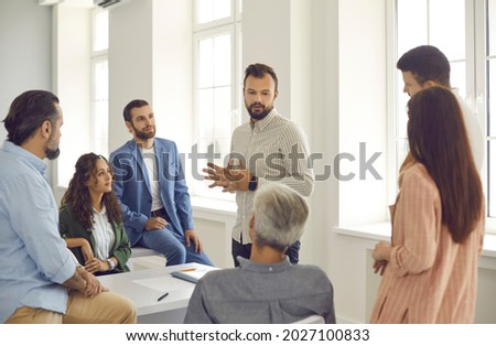 Small group of diverse women and men listen to a confident male corporate business coach. Man advises clients, trains employees or explains the strategy of a marketing seminar. Staff training concept. Royalty-Free Stock Photo #2027100833