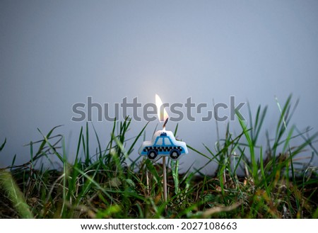 A festive candle in the form of a blue car, burning with a bright flame on a white background, green grass at the bottom, a place for a greeting text