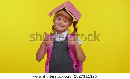 Smiling cute schoolgirl wears backpack, reading book, making playful silly facial expressions and grimacing, fooling around, putting book on head over yellow studio background. Back to school concept