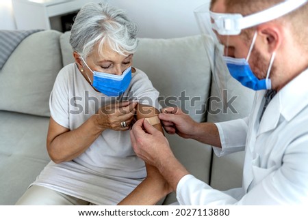 A brave senior woman sitting with her pediatrician at the office as she receives a bandage after a vaccination. Beautiful old female patient receiving a bandaid from a young male Caucasian physician.