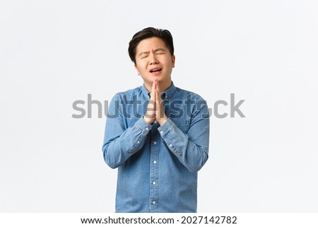 Overworked gloomy and sad asian man begging for help, holding hands together over chest in praying gesture, asking favour, showing remorse, standing white background overwhelmed Royalty-Free Stock Photo #2027142782