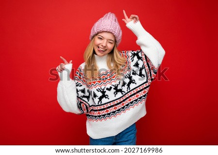 Photo of young positive happy attractive blonde woman with sincere emotions wearing pink knitted hat and winter sweater isolated over red background with free space and dancing