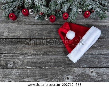 Snow covered evergreen branches with bright red burning candles and a Santa cap, on faded wooden planks for a merry Christmas or happy New Year holiday celebration concept 